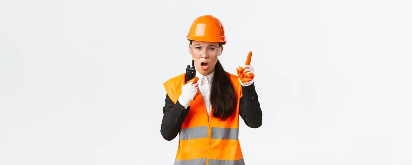 Ernst dreinblickende asiatische Industrieingenieurin und Technikerin mit Schutzhelm und Uniform an einer Steuerkonsole.