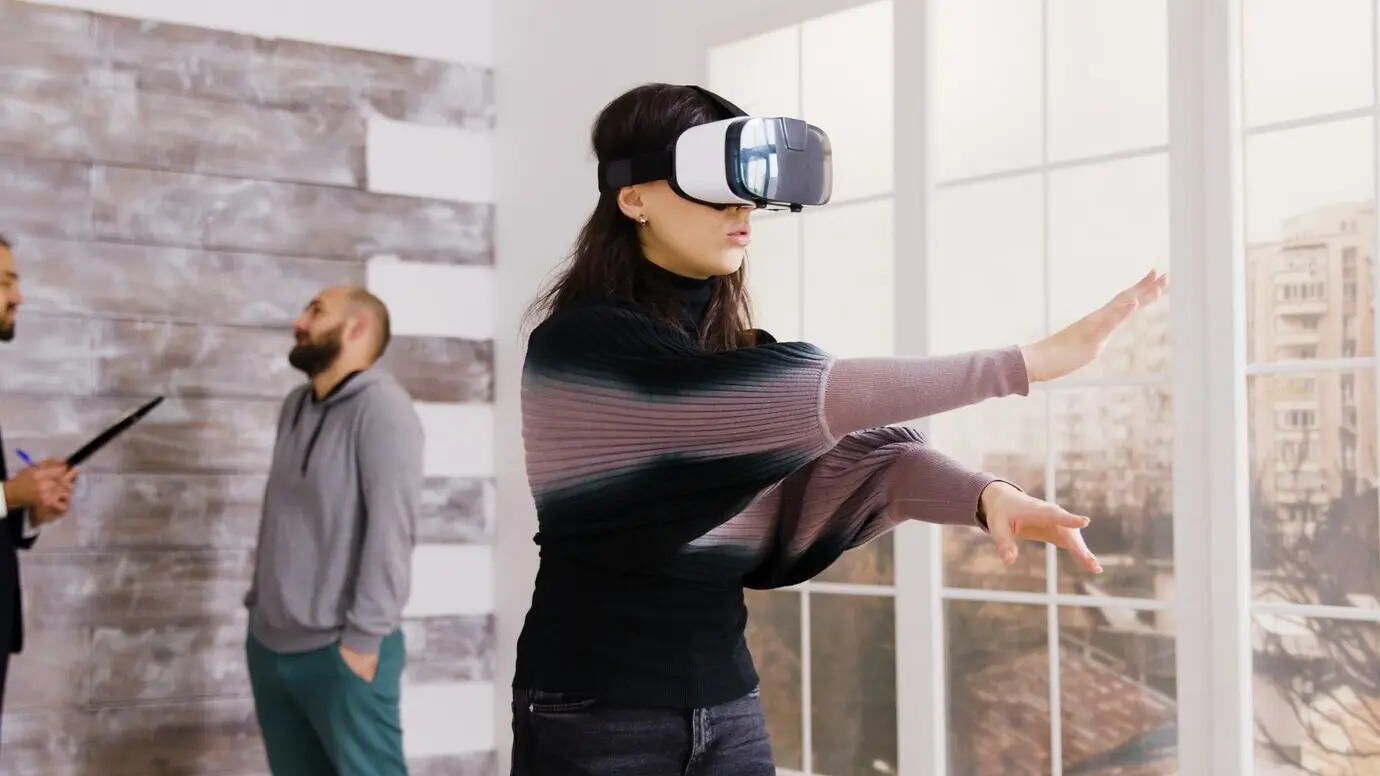 Eine Architektin nutzt in einer leeren Wohnung eine Virtual-Reality-Brille, während im Hintergrund ein Immobilienmakler mit einem Kunden spricht.